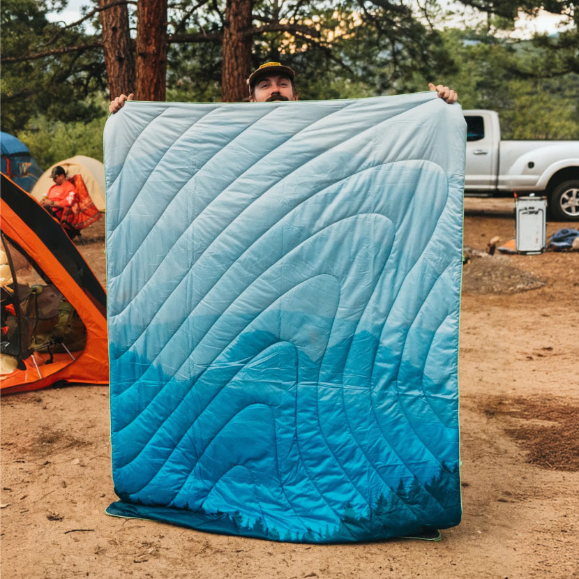 Person holding a blue gradient sleeping bag outdoors near tents.