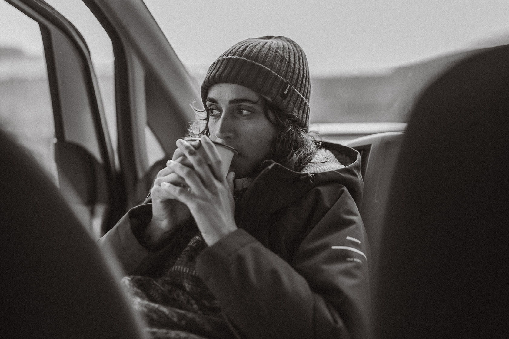 Person sitting in a car, drinking from a cup, wearing a beanie.