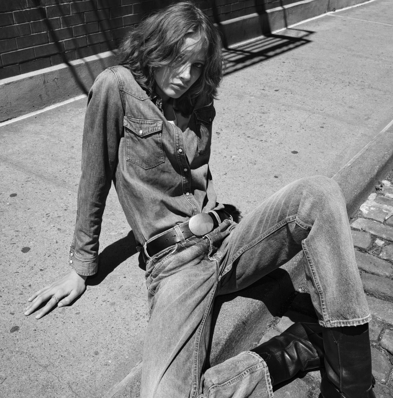 Person sitting on a sidewalk wearing a denim outfit and boots.