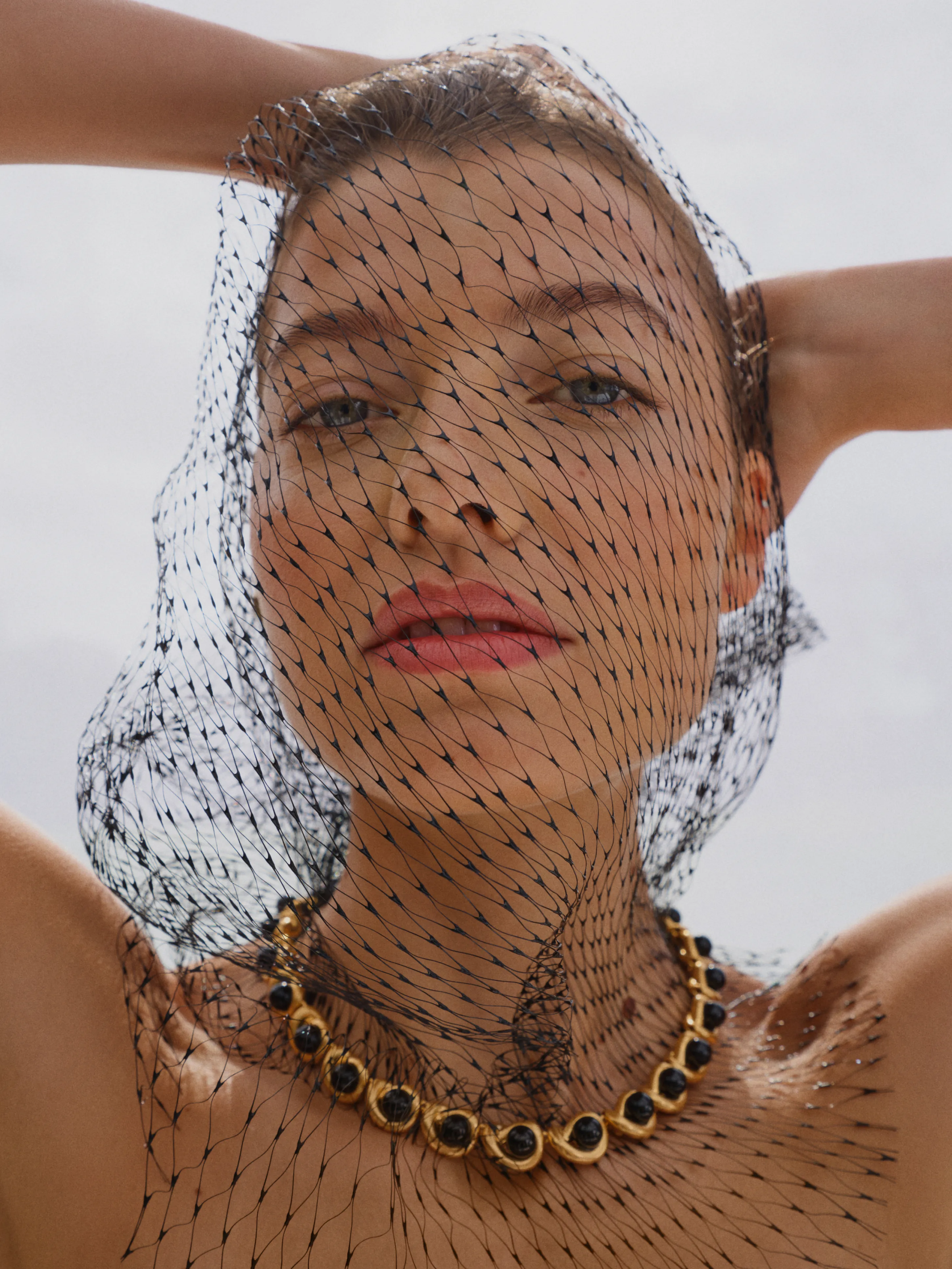 Woman with a netted veil and necklace, posing against a light background.
