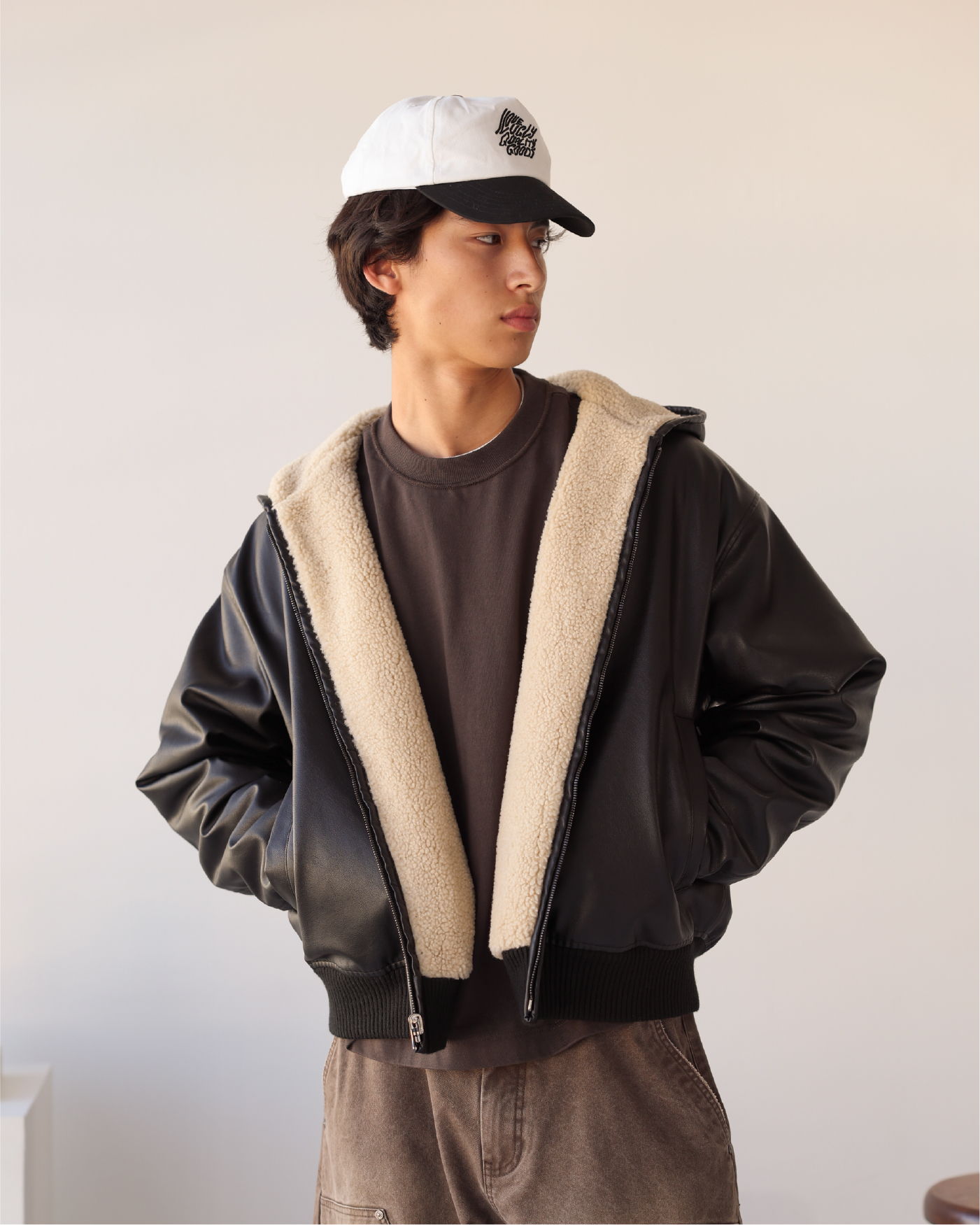 Young man in a black jacket and white cap, standing against a neutral background.