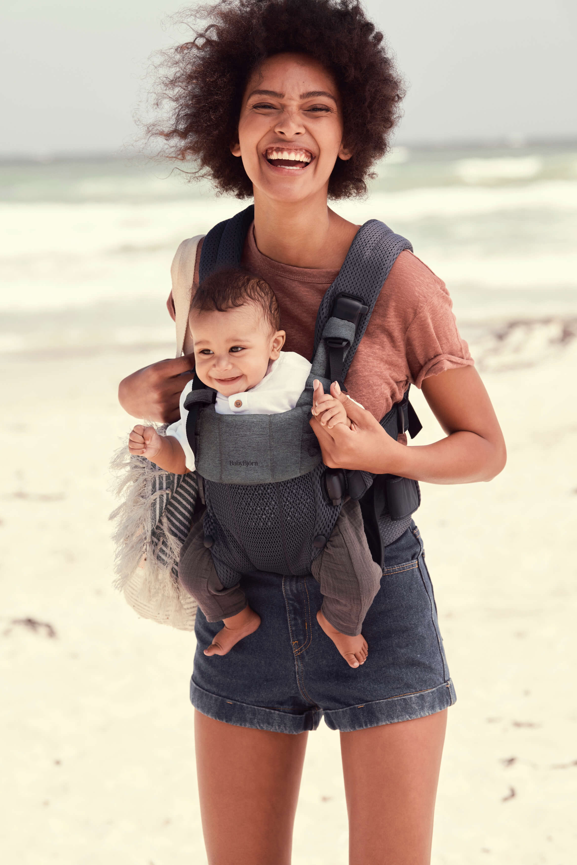 Smiling woman at the beach carrying a baby in a front-facing carrier.