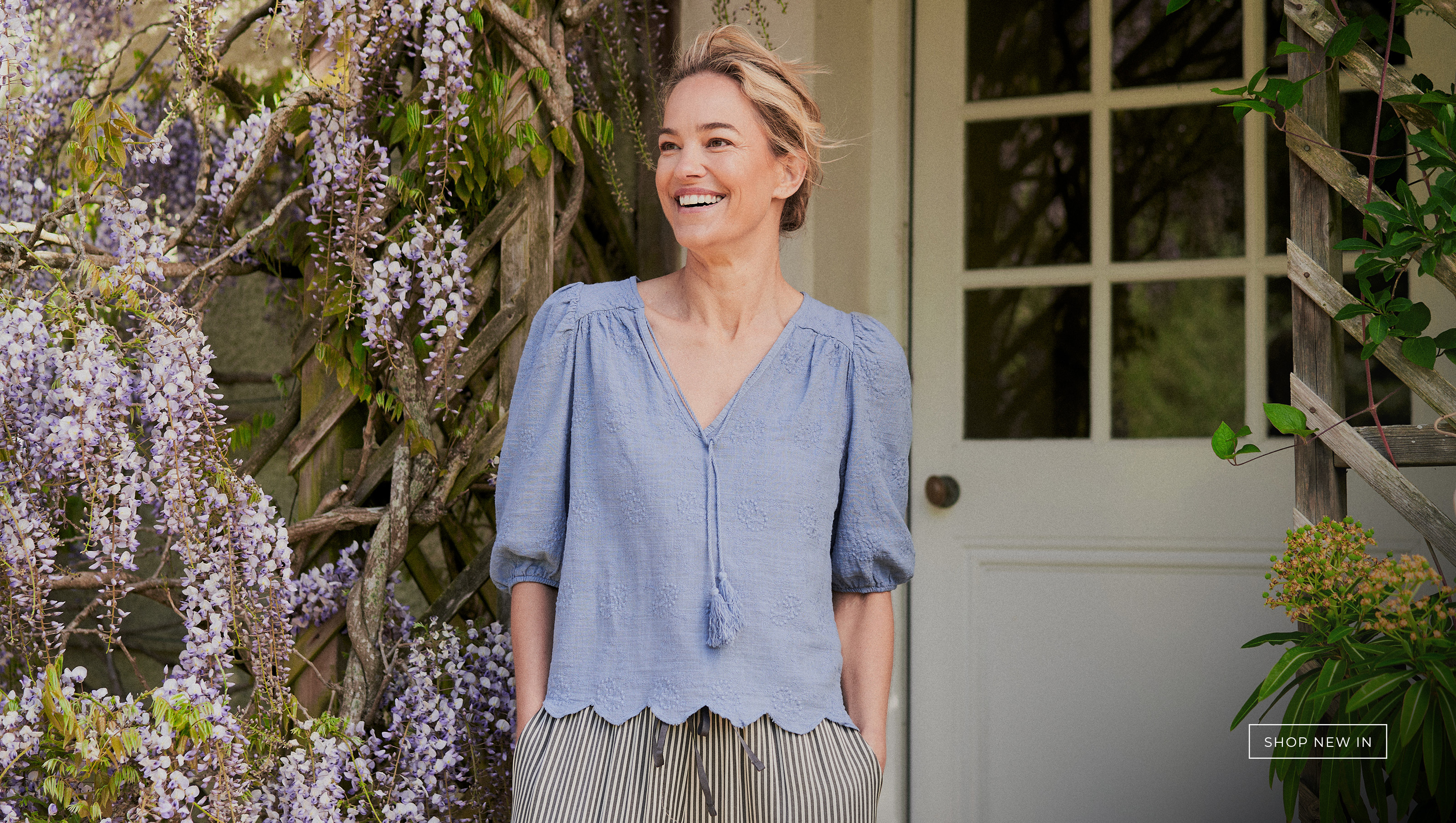 A woman standing in a doorway surrounded by wisteria flowers in a light blue embroidered top