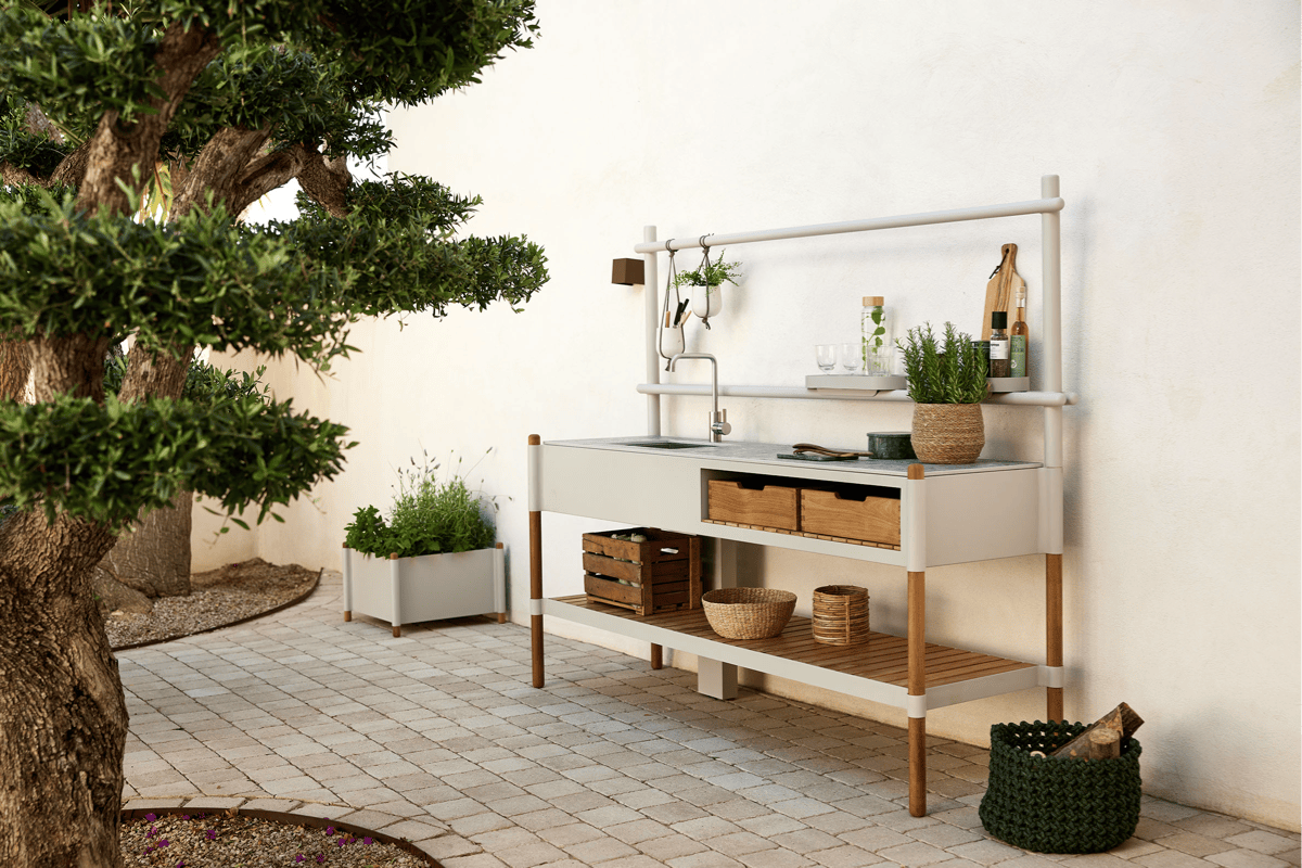 Outdoor prep station with sink and open shelving against a white wall, styled with bottles, herbs, and storage baskets.