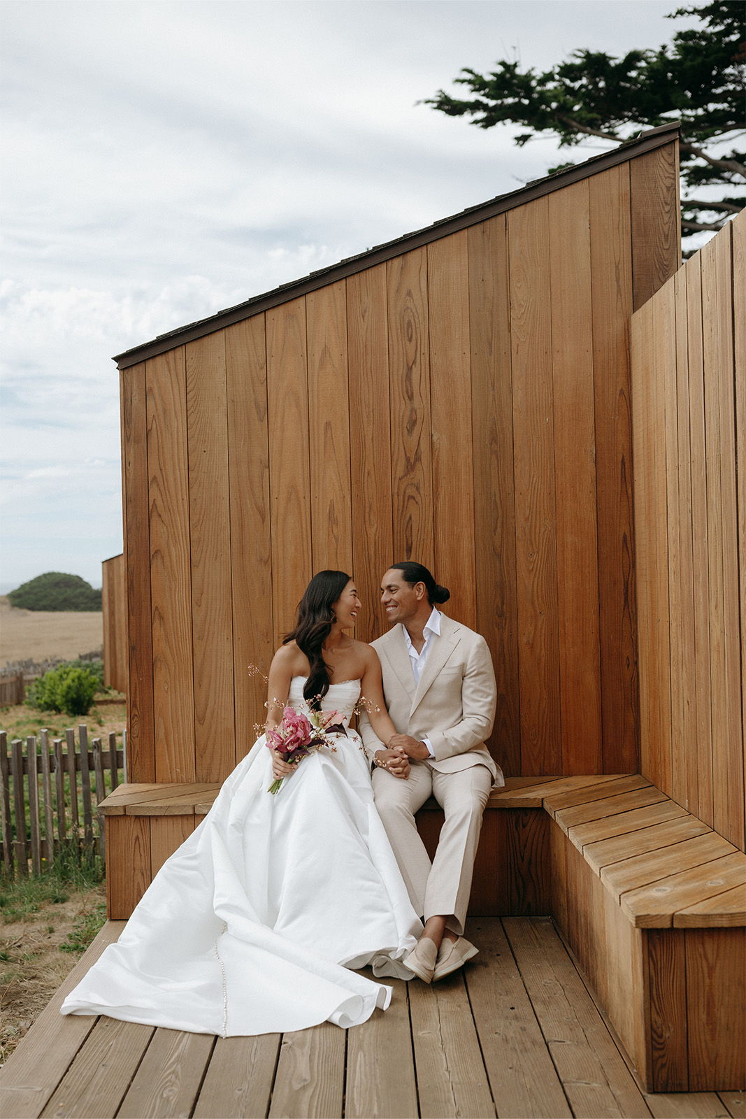 Bride and groom sitting together on a wooden bench, smiling and holding hands.