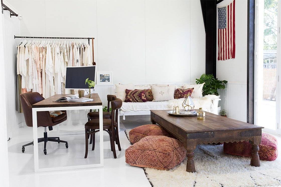 A light white office space featuring a desk with computer, coffee table with floor pillows, a white couch and a rack of clothes. 