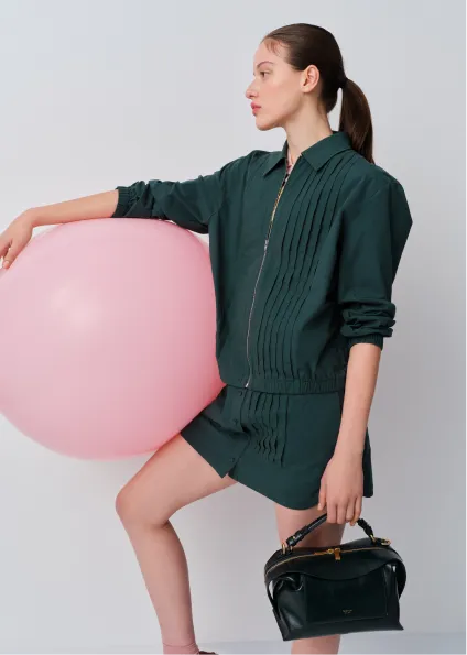 Model in green outfit holds pink balloon and black handbag.