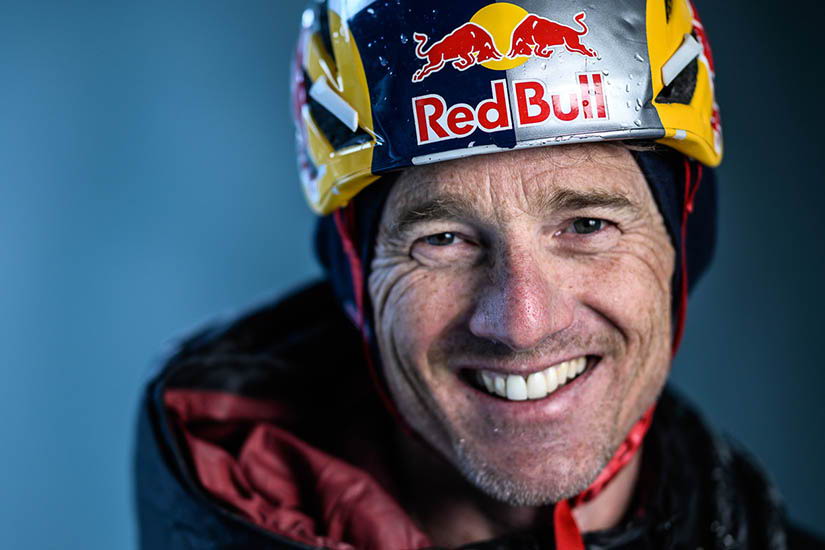 Smiling athlete Will Gadd wearing a helmet with Red Bull logo against a blue background.