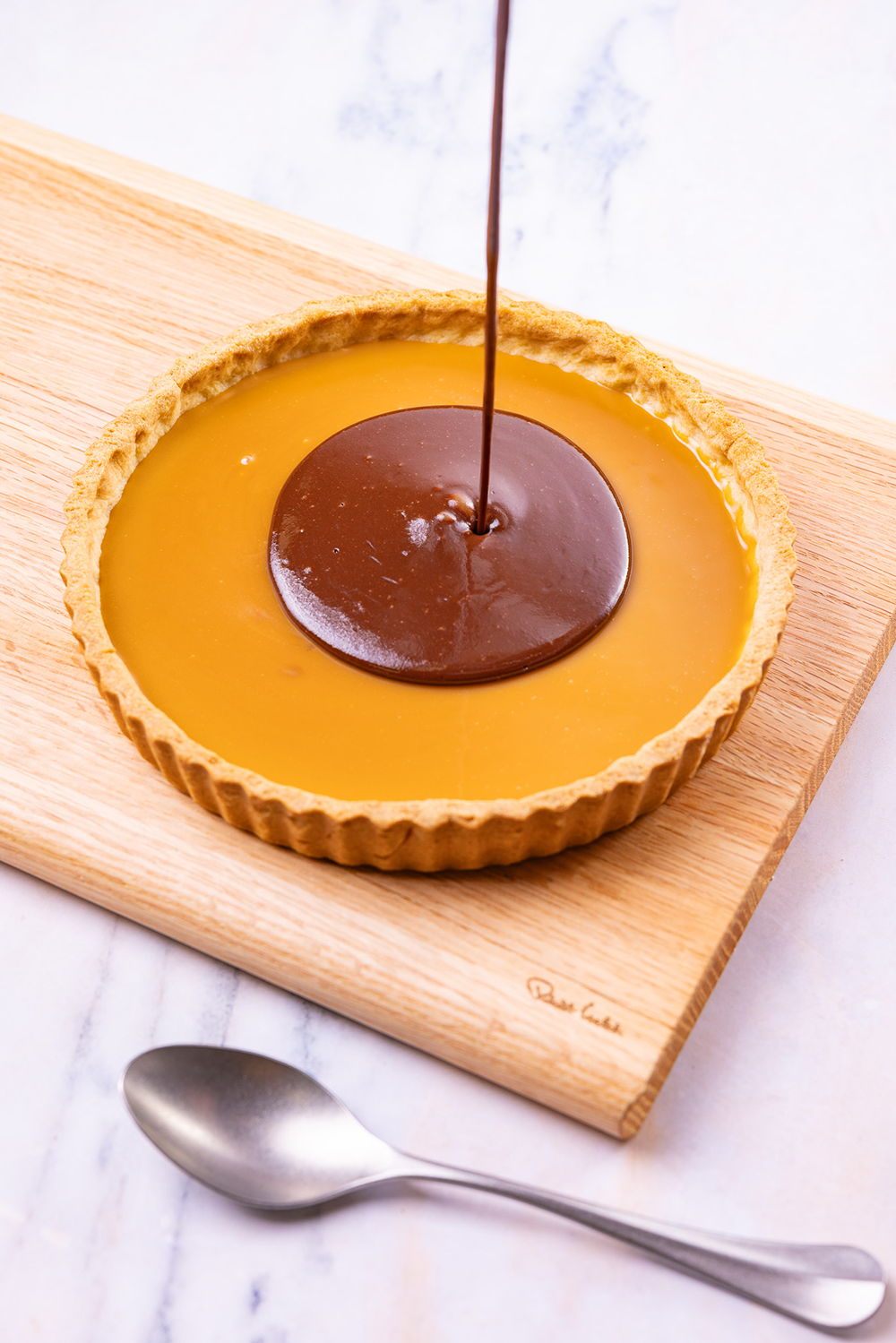 Chocolate sauce being poured onto a caramel tart on a wooden board.