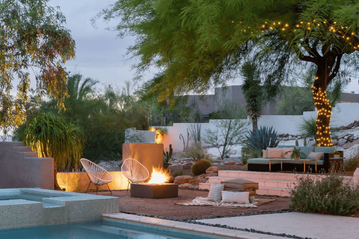 A poolside fire pit area illuminated t night featuring chairs, outdoor rug, and throw pillows. 