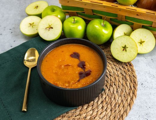 Bowl of butternut squash and strawberry soup with green apples and a golden spoon on a table.