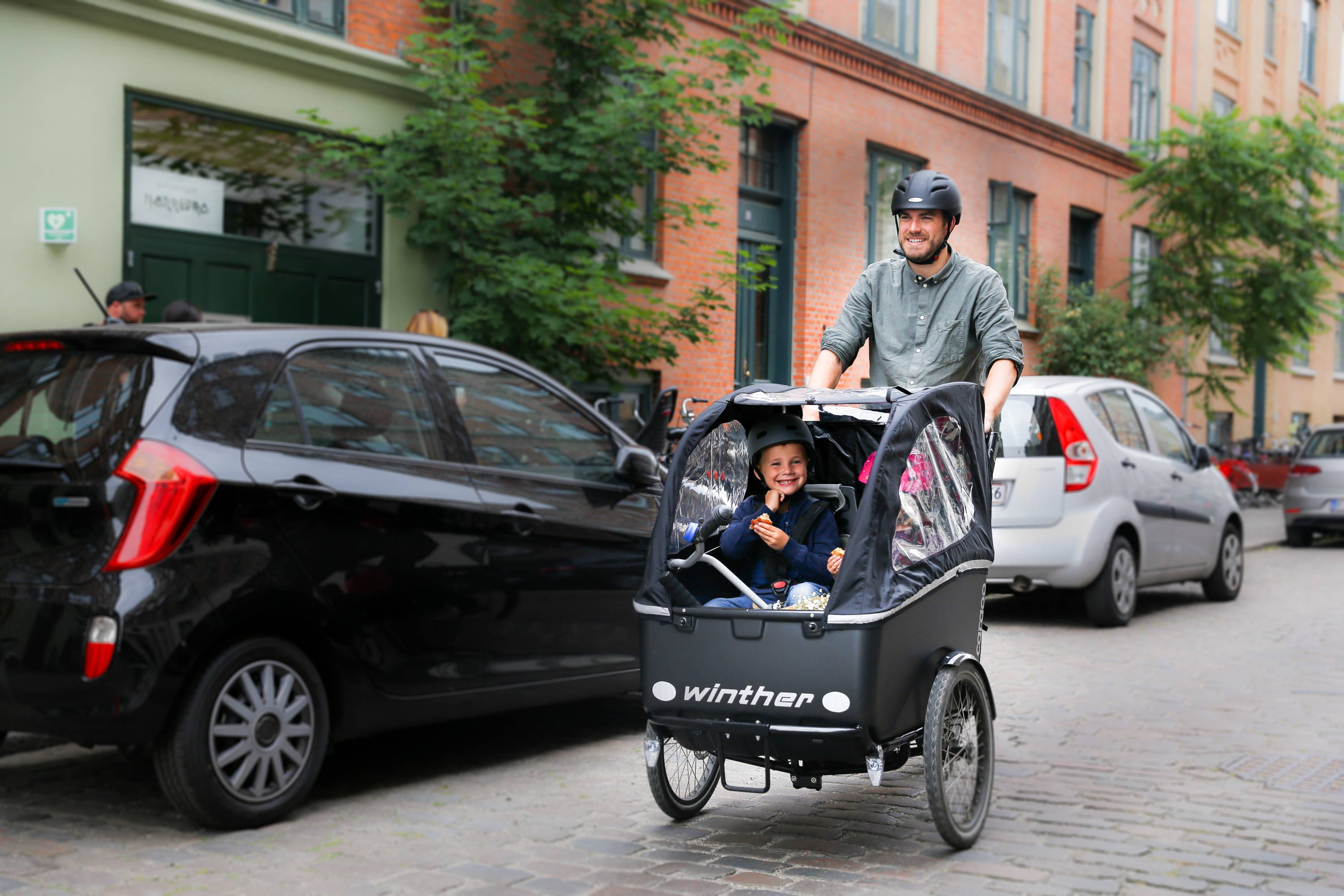 A man rides a Winther cargo bike through a European side-street with a smiling child in the front box.