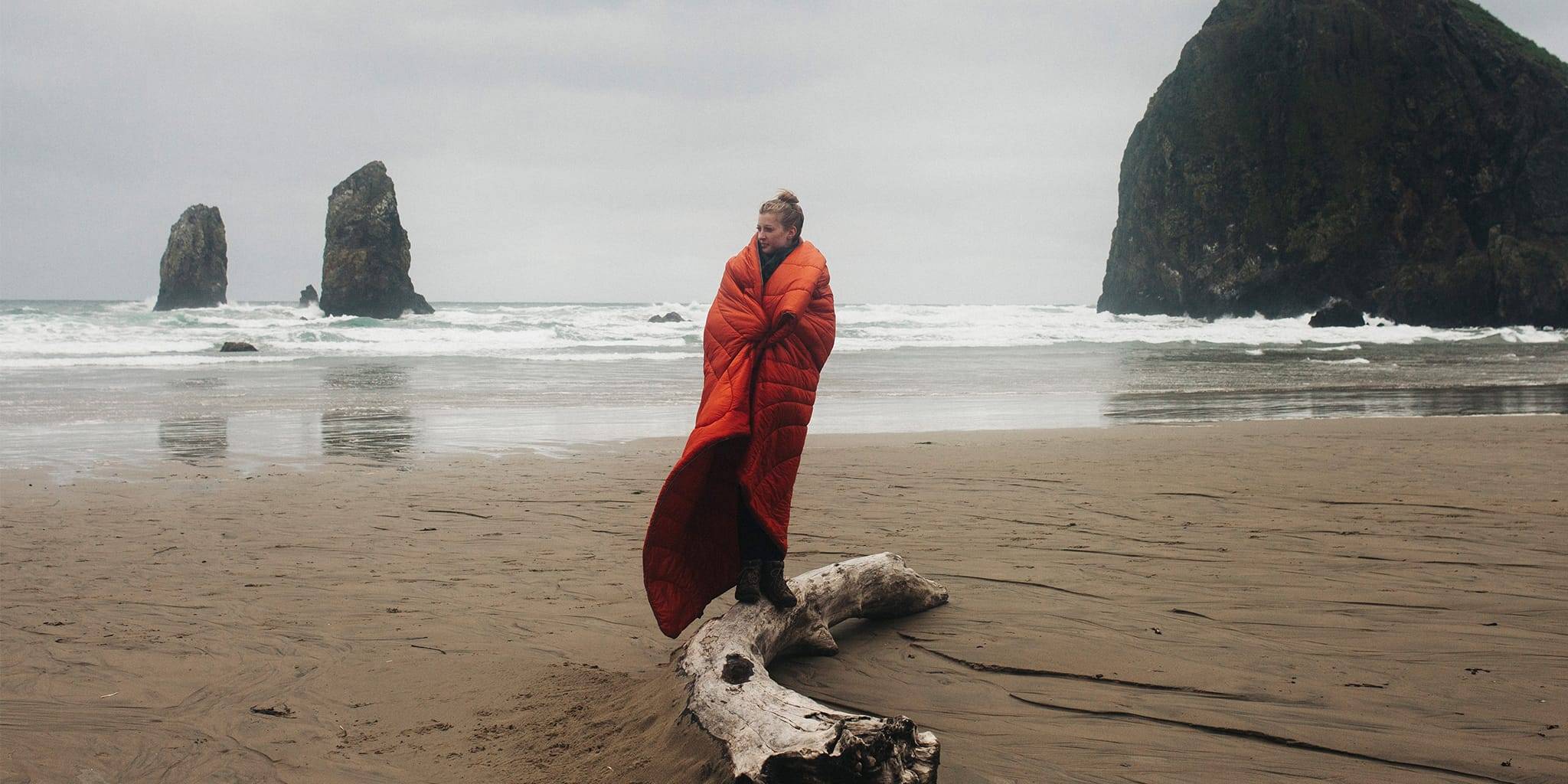 Kristian Irey standing on driftwood by Haystack Rock with an Orange Rumpl blanket