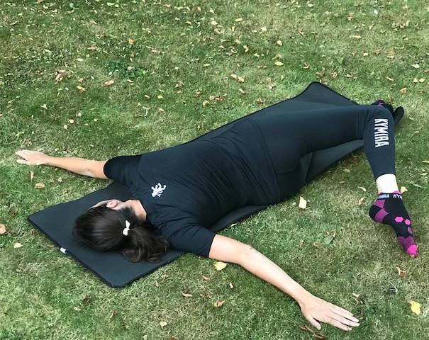 A woman in KYMIRA gear lies on the floor in a T formation, she rolls her body to the right and has her left heel across the body.