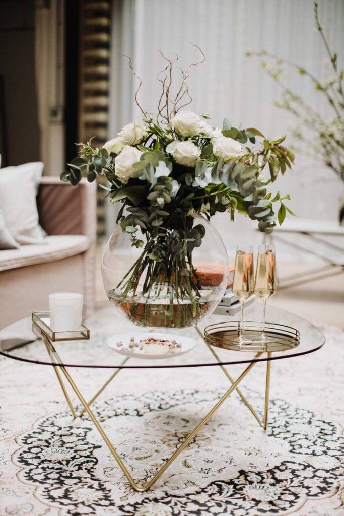 White roses in a clear round vase with champagne glasses on traditional rug