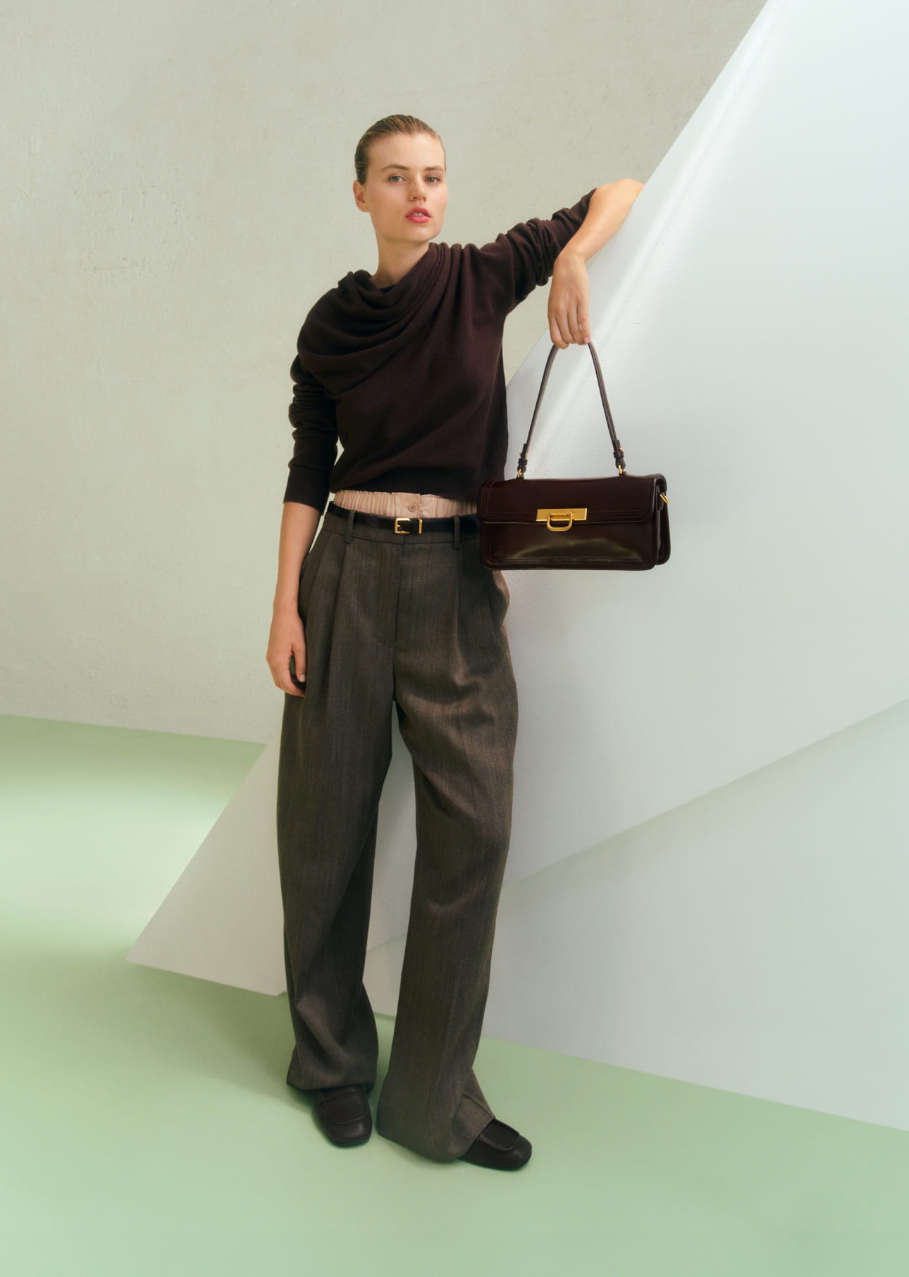 Model in a black sweater and wide pants, holding a handbag against geometric backdrop.