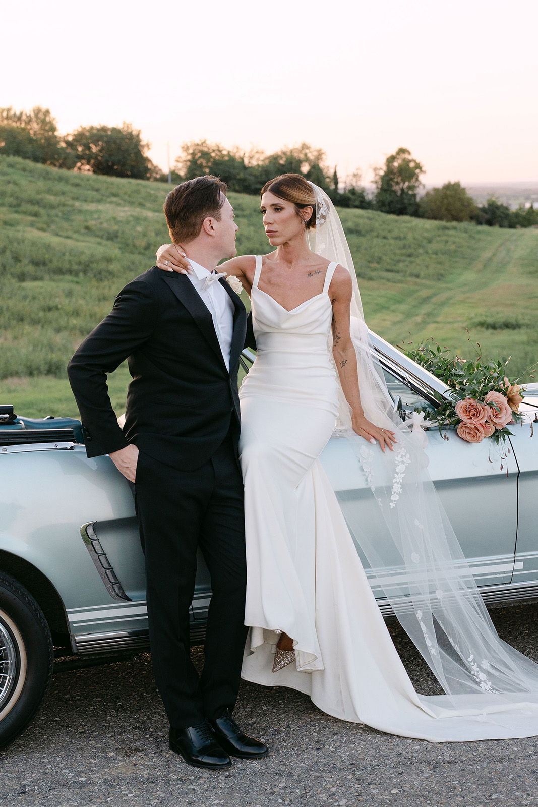 Bride and groom posing by a vintage car in a scenic outdoor setting.