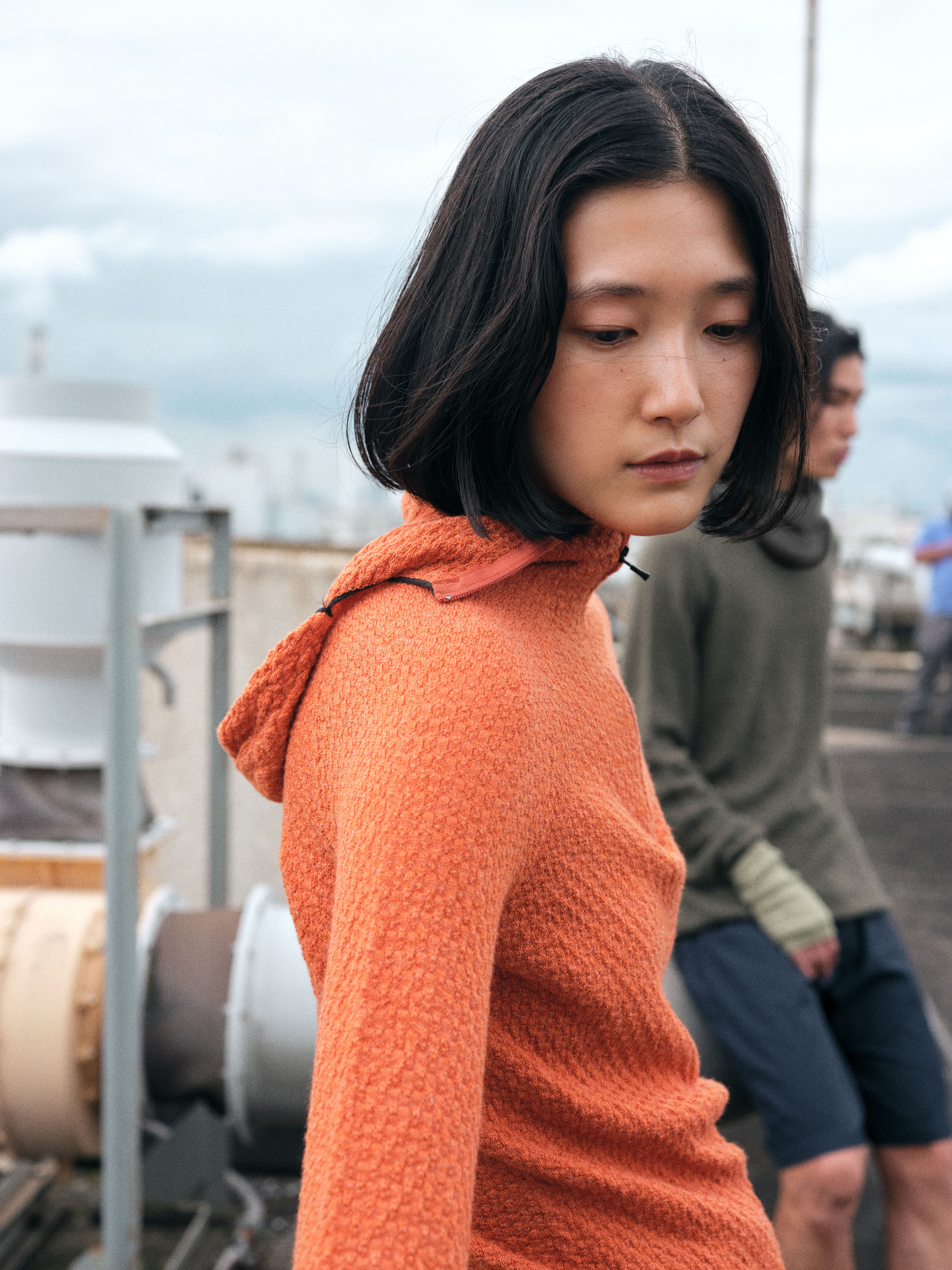 Person with bobbed hair wearing an orange sweater, outdoors with cloudy background.