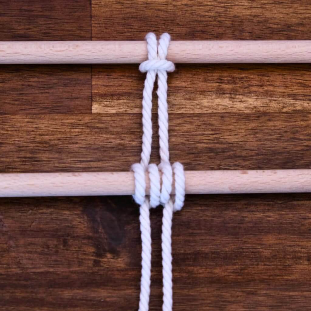 Zwei Holzstäbe, zwischen denen ein weißes Seil zu einem Rippenknoten gebunden ist.