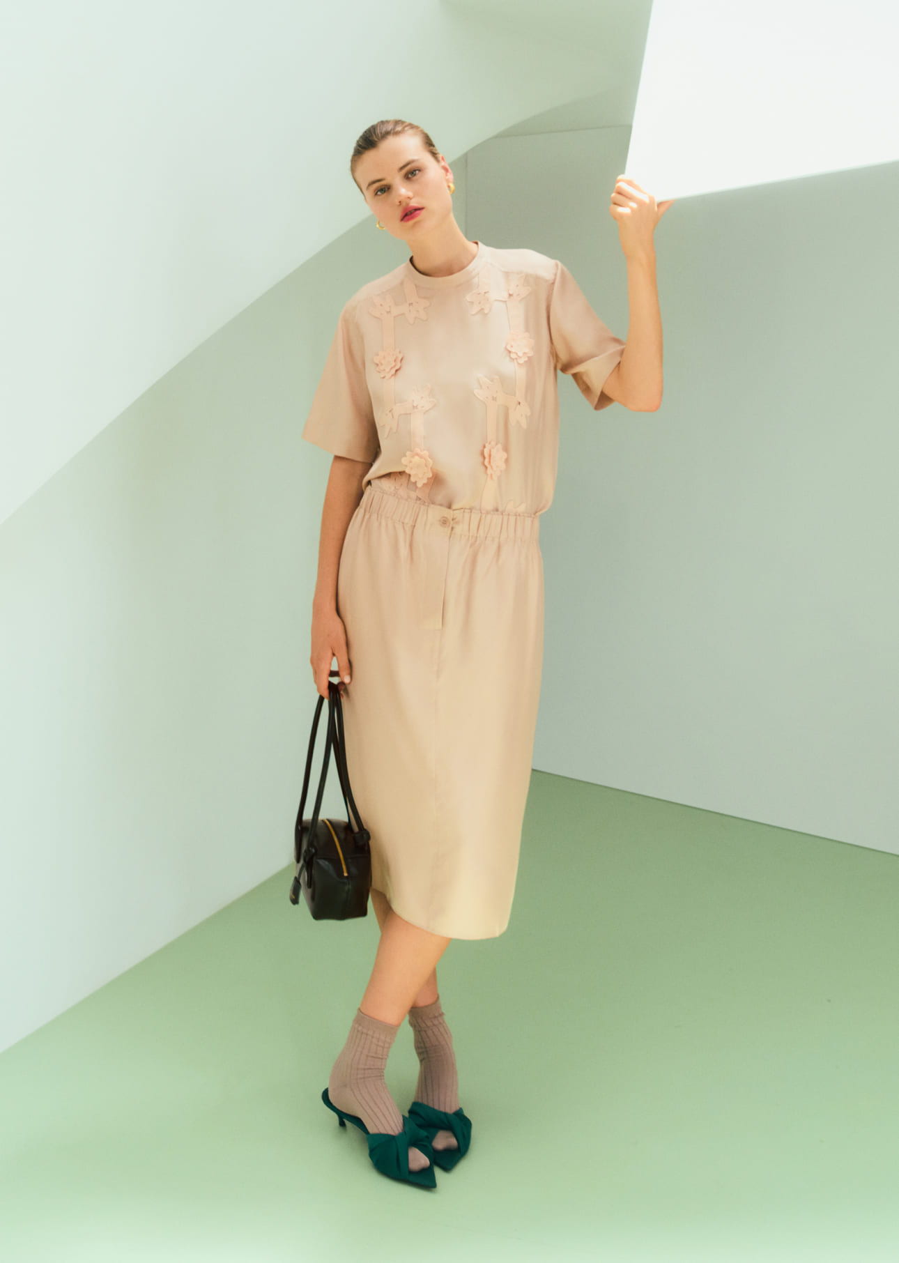 Model in a beige dress and blue shoes, standing against a pastel background.