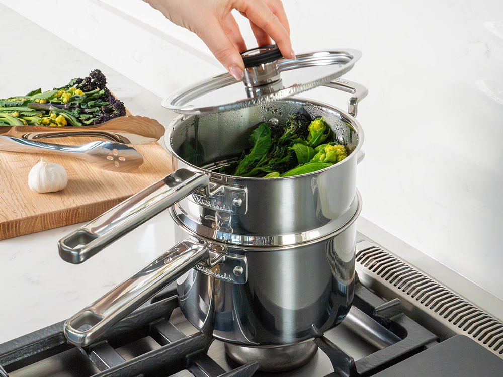 Hand lifting the lid of 316 Series steamer pot with broccoli on the stove.