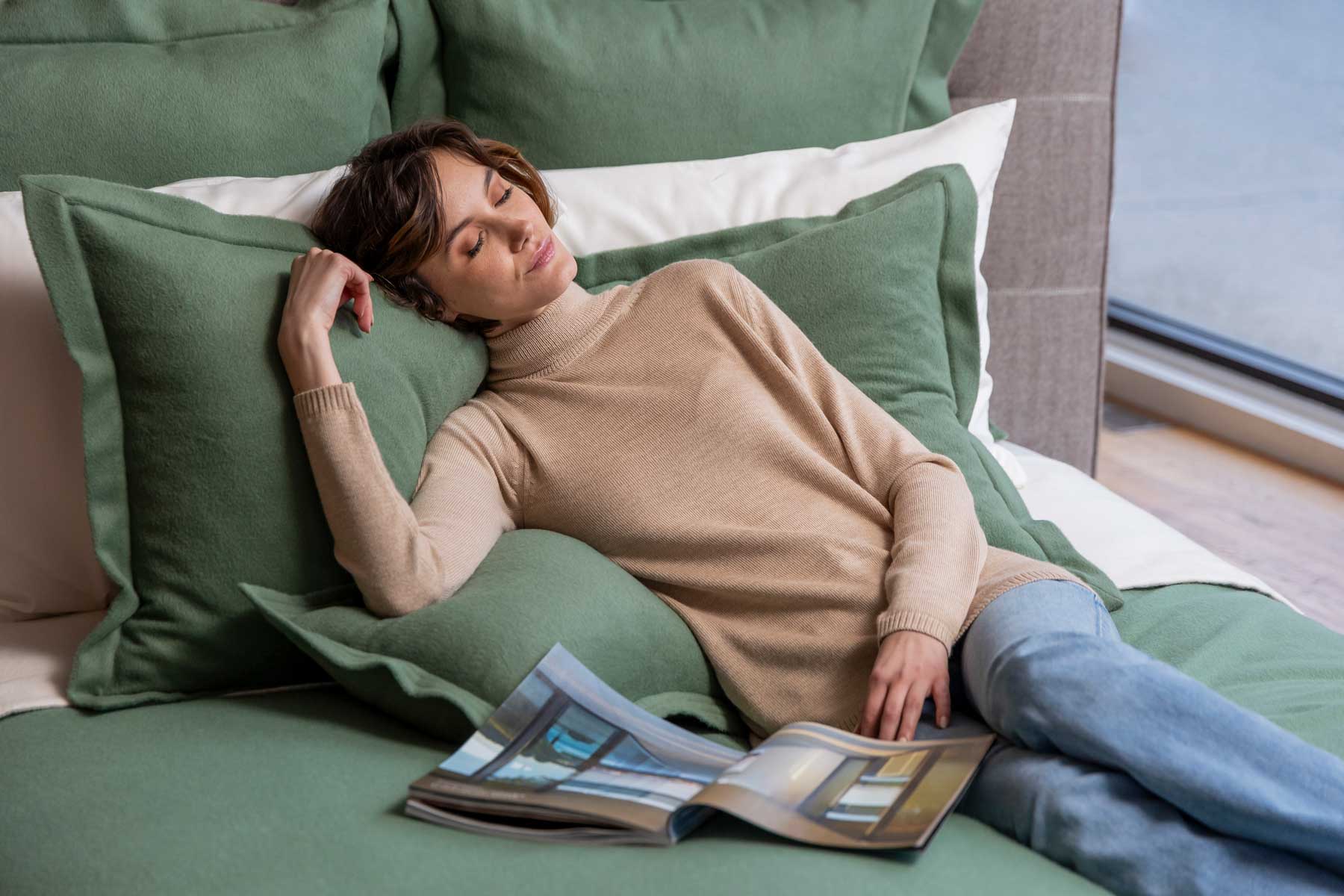 Person lounging on a bed with green pillows, holding a magazine.
