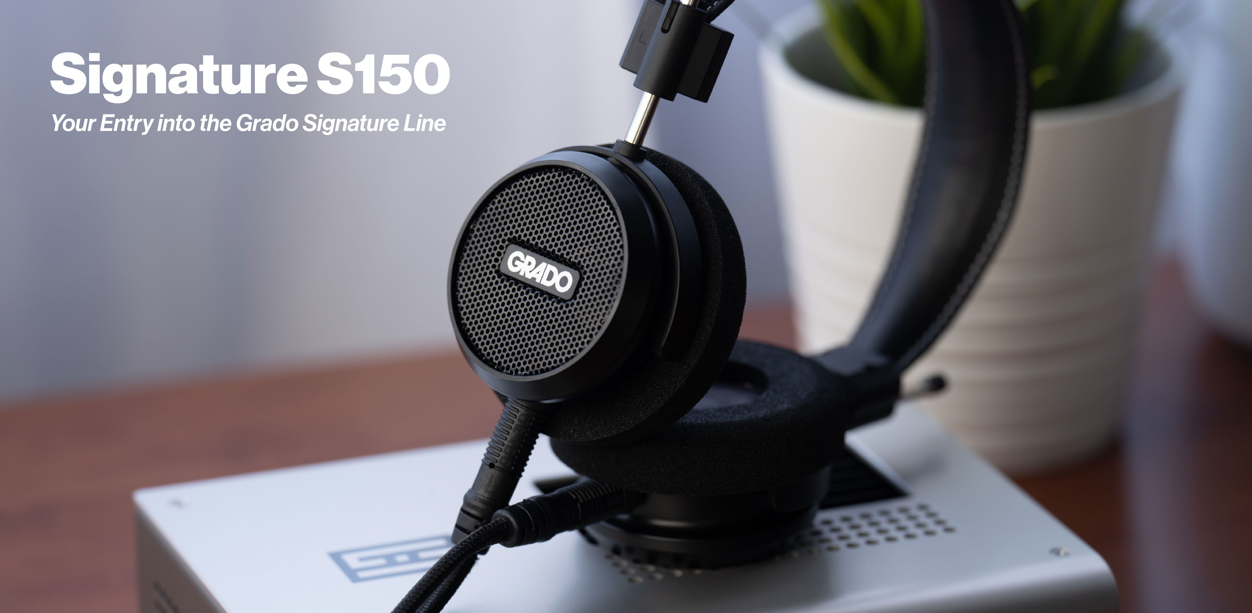 A photo of the S150 headphones resting on one ear cup on a wooden table. There is text in the top left corner that says 'Signature S150' with 'your entry into the grado signature line' underneath.