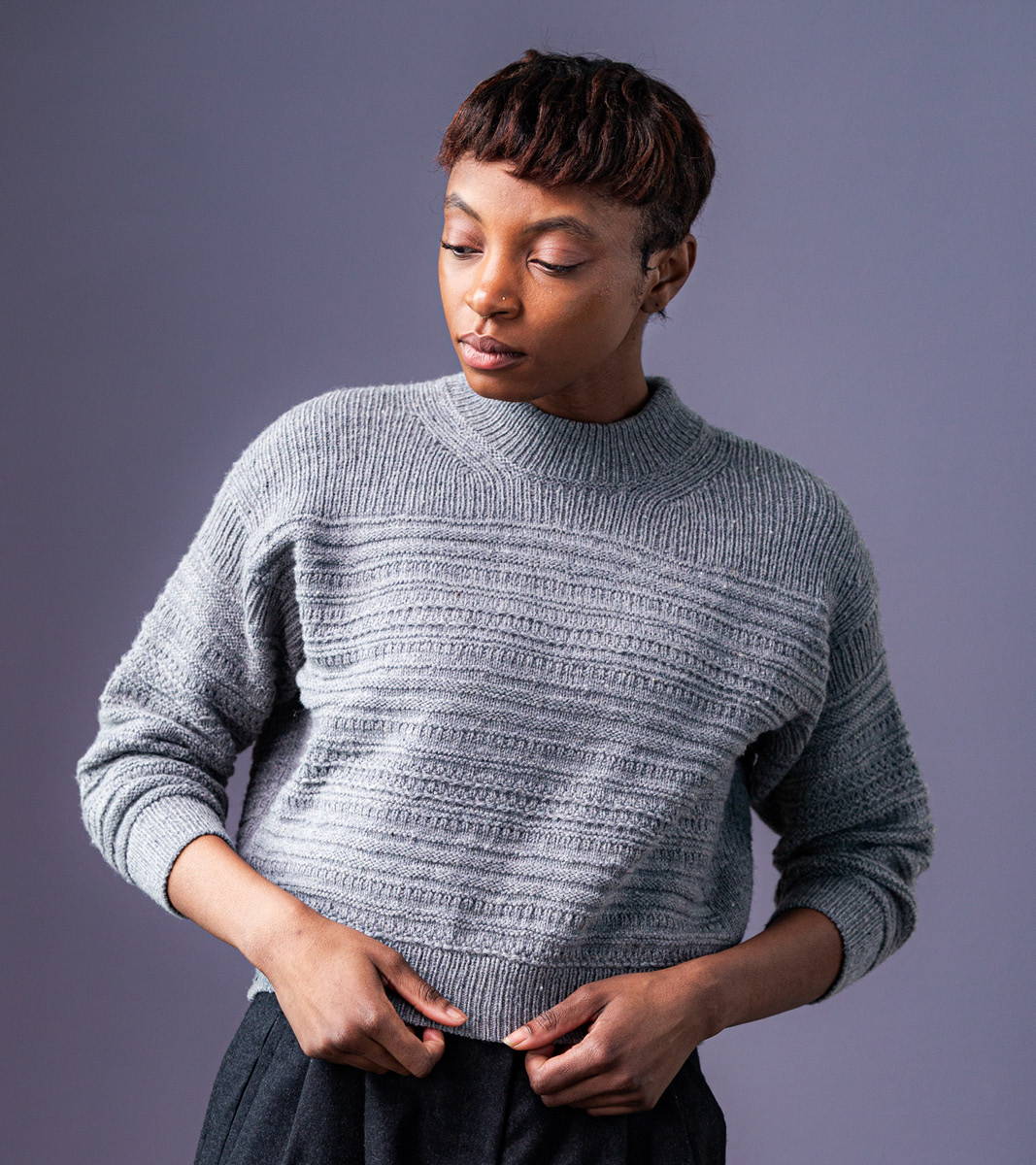 Maya, a woman with short brown hair, models Brooklyn Tweed's Grist Pullover hand knit in Imbue Sport yarn color Ash