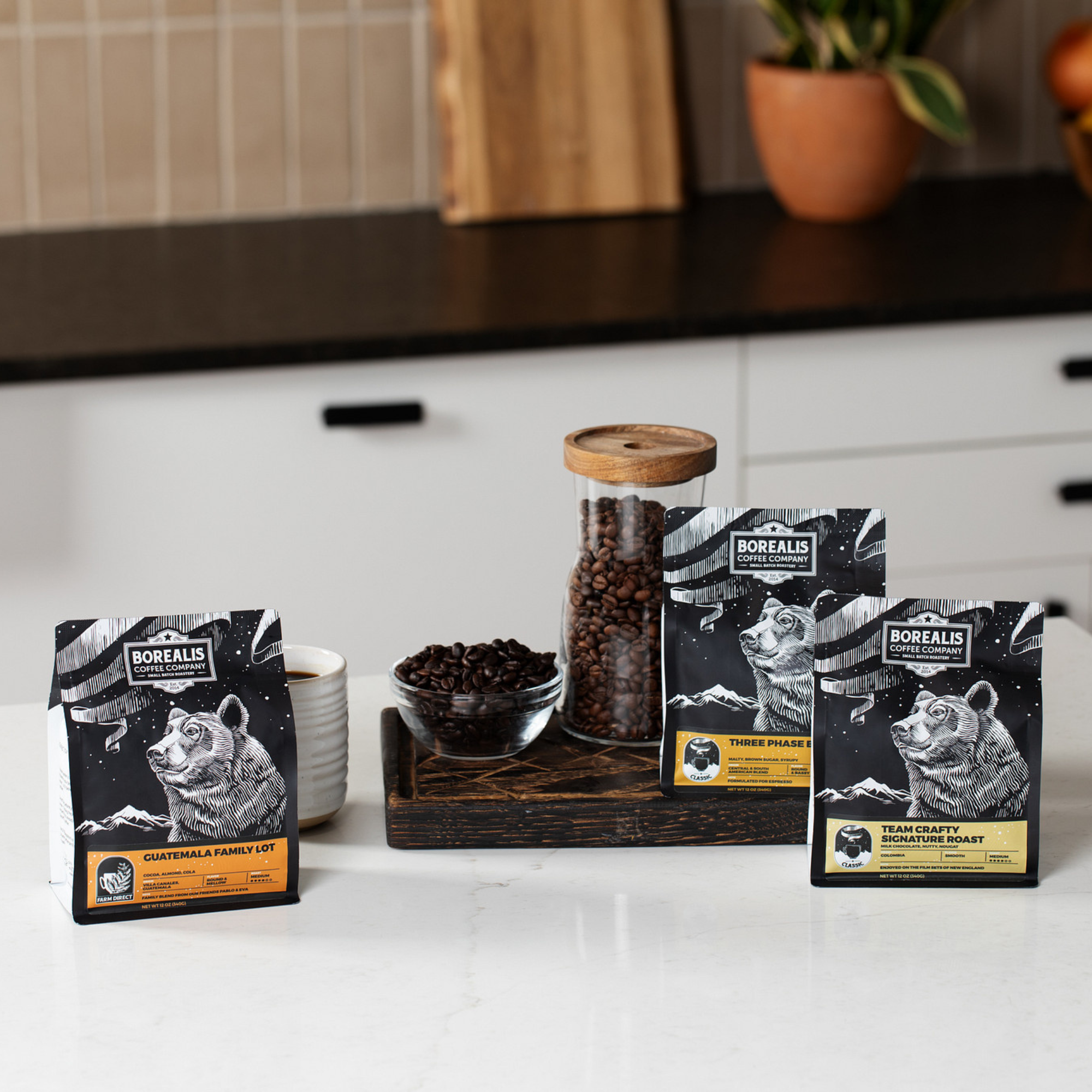 Three bags of Borealis coffee staged on a white kitchen counter with a cup of coffee and glass jar of coffee beans to illustrate a typical subscription box.