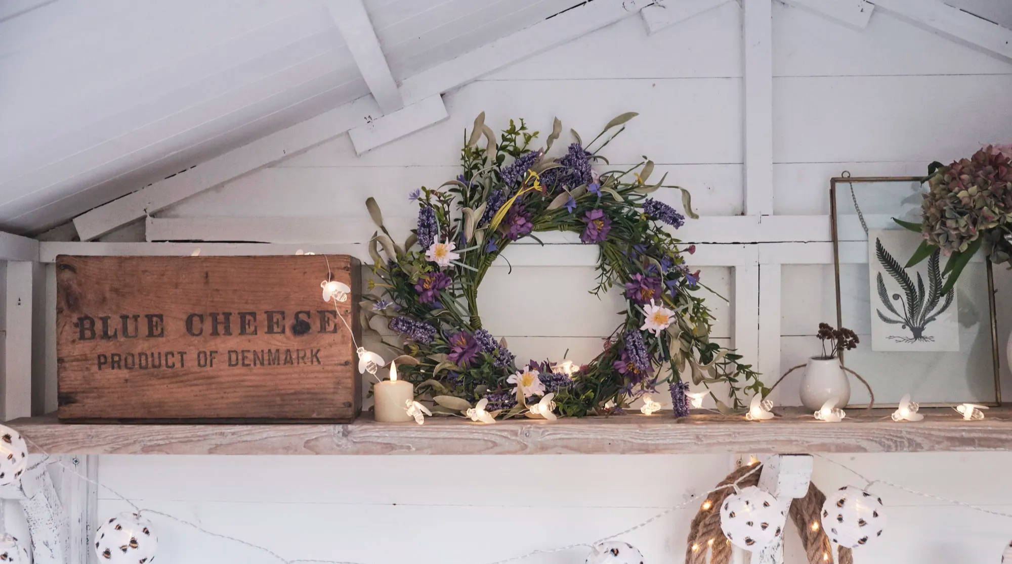 A lavender spring wreath on a shelf in a summer house surrounded by bee lights, LED candles and paper lights.
