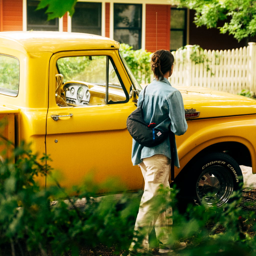 Person standing by a yellow vintage truck in a green, leafy setting.