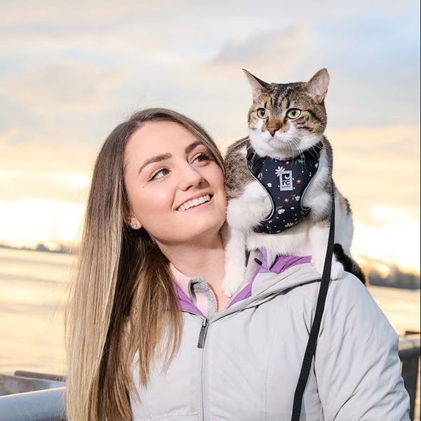 A woman smiles with a cat on her shoulder against a sunset background.