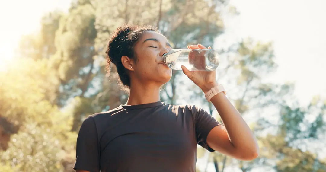 Person drinking water outdoors surrounded by trees in bright sunlight.