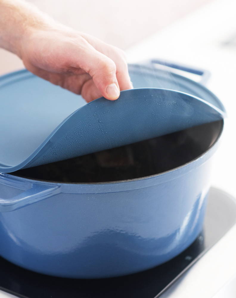 A hand peeling a blue silicone lid partially off a blue Misen Dutch Oven to allow steam to escape.
