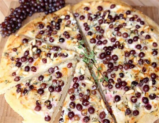 A pizza topped with grapes and herbs, sliced on a wooden board.