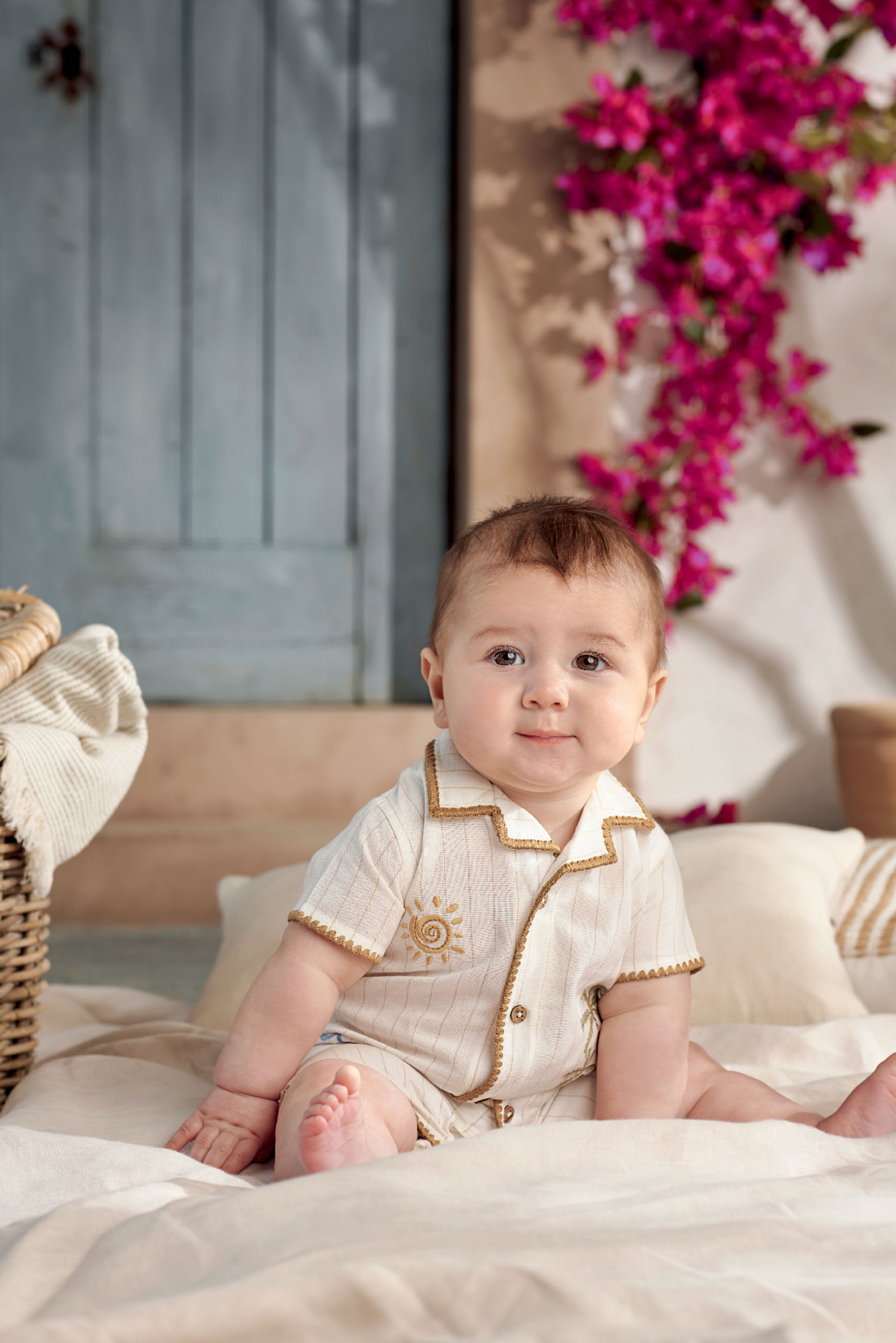 A baby sitting on a bed surrounded by flowers and a blue door.