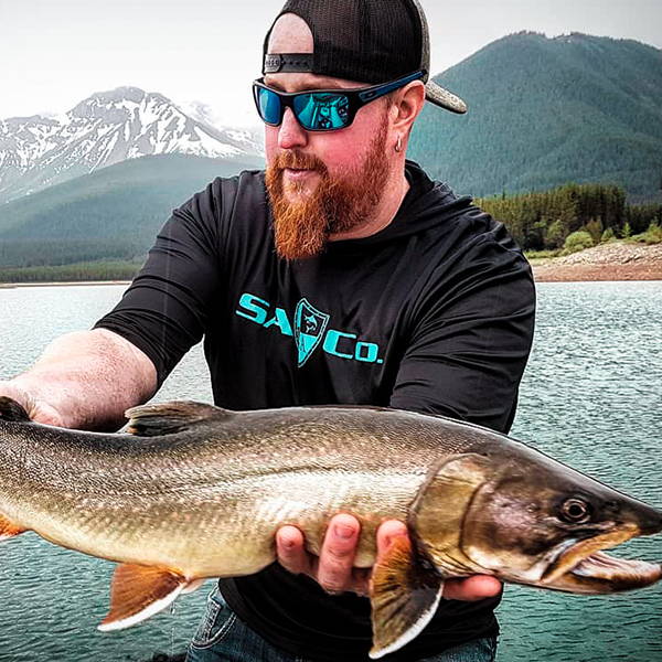 Mitch Dupuis by a lake holding up a fish to the camera while wearing an SA Company black cotton shirt, sunglasses and a hat.