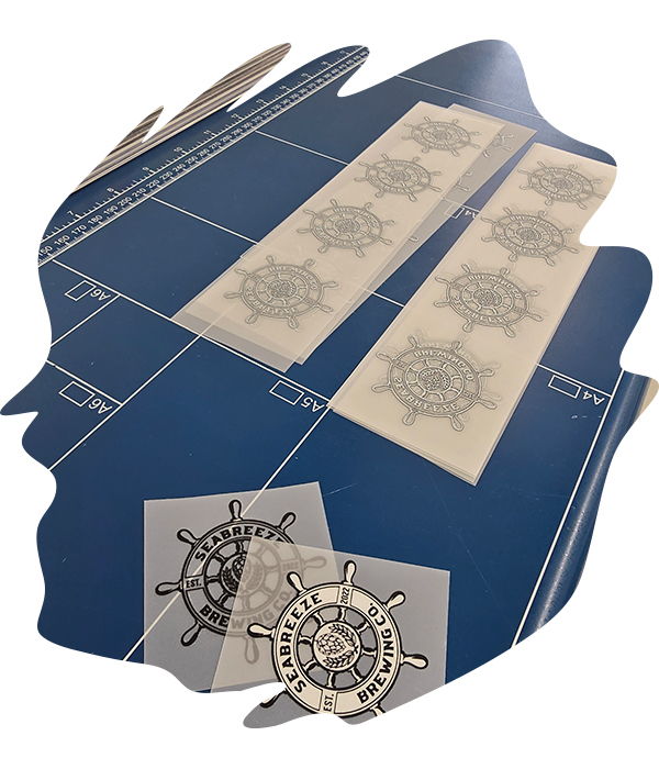 Sheets of multiple cured Seabreeze Brewing Co. ship wheel DTF transfers laid out on a blue cutting mat, with two individually cut transfers placed beside it