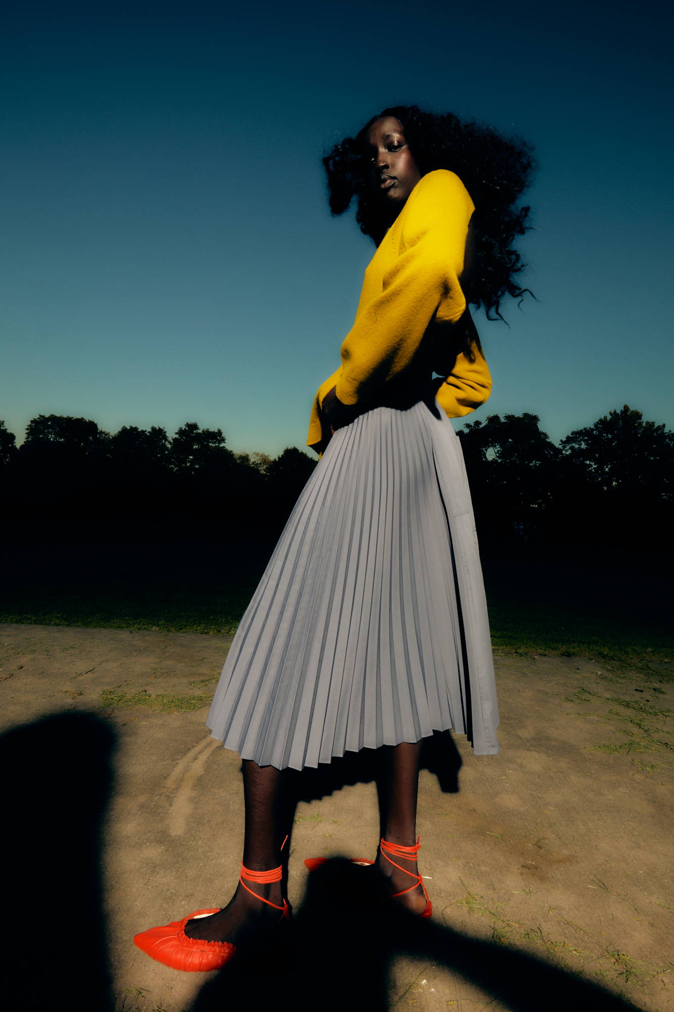 Model in a yellow sweater and grey skirt, posing outdoors at sunset.