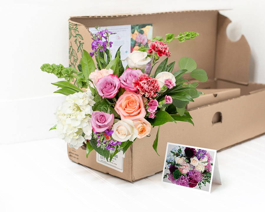 A box holds the Enjoy Flowers Farm Fresh collection of pink, white, and purple roses with foliage and a personalized card. 
