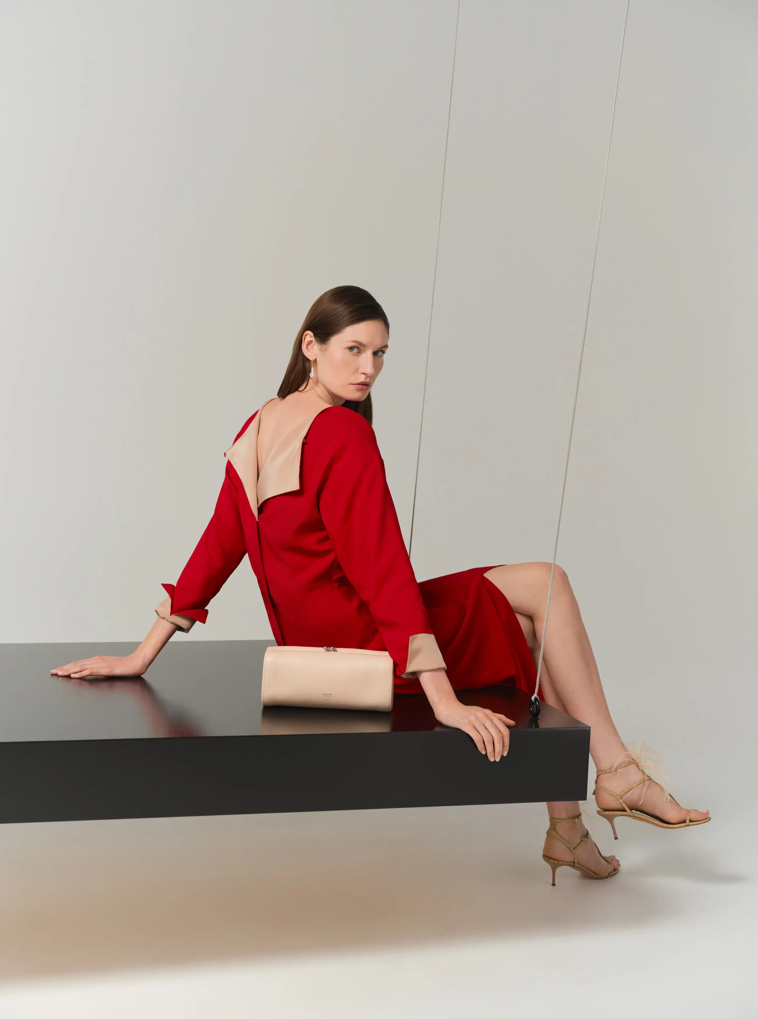 Model in a red dress seated on a black surface, holding a clutch.