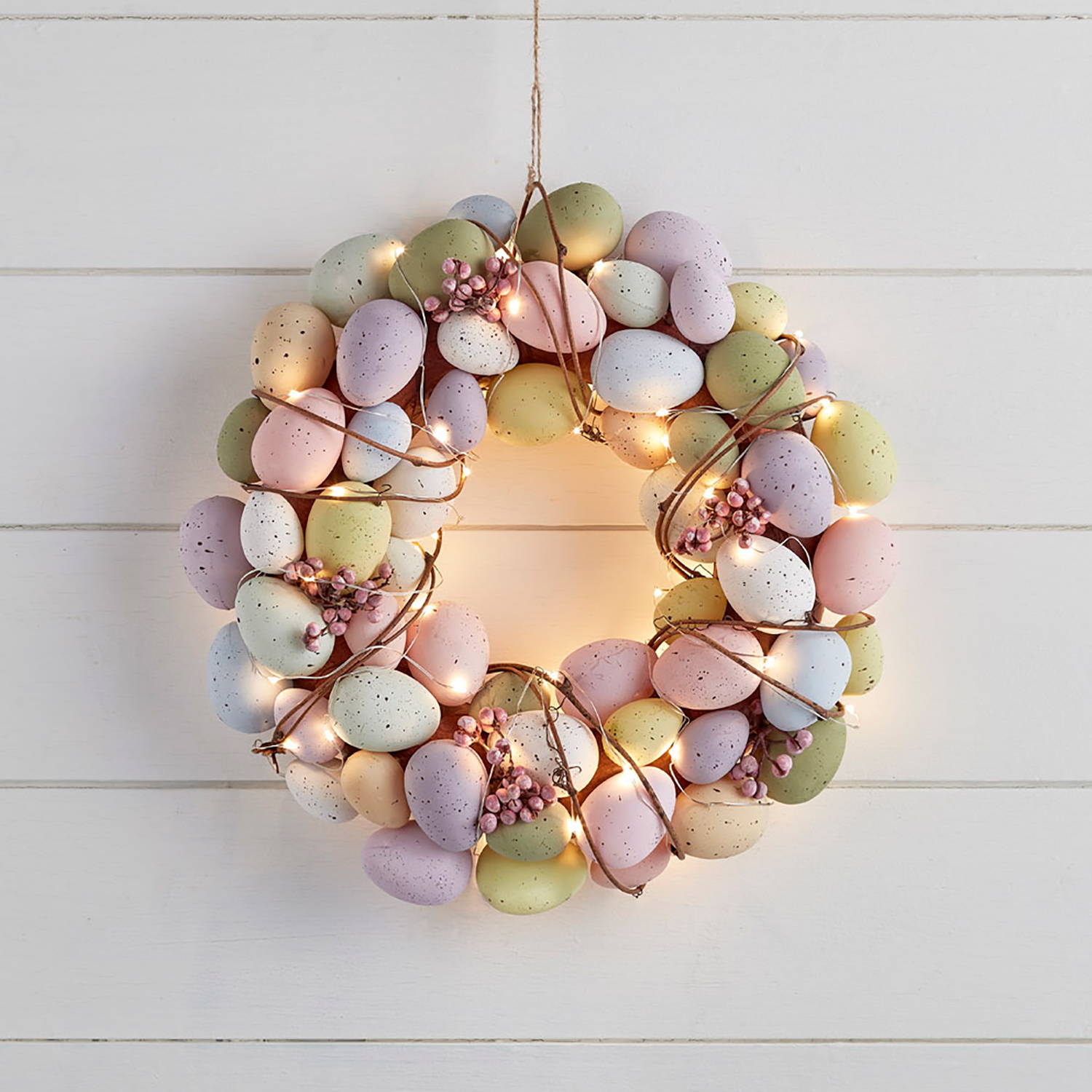 An multicoloured pastel egg round garland with berries and micro lights against a white wall.