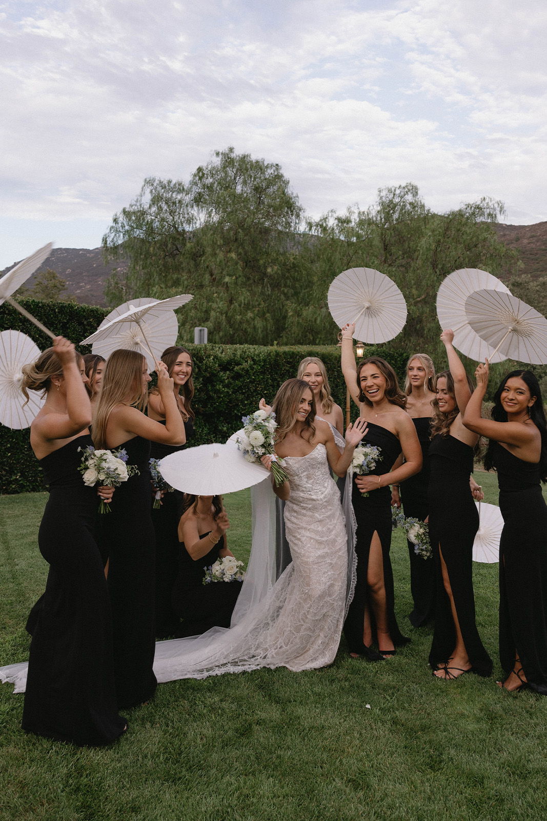 Bride and bridesmaids celebrating outdoors, holding fans and flowers, wearing elegant dresses.
