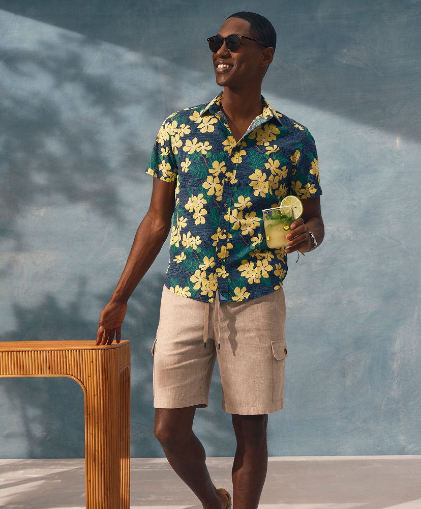 Man in floral shirt holding a drink, standing next to a wooden table.