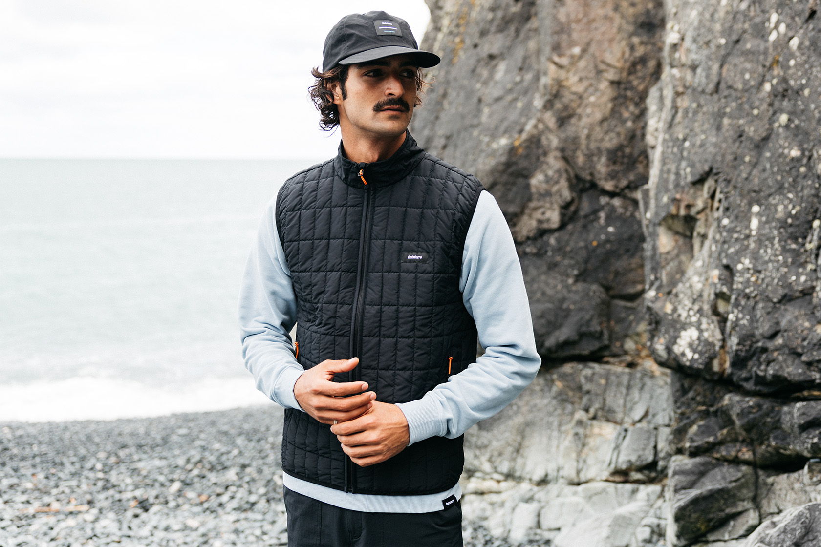 Man in a vest and cap stands by rocky beach.