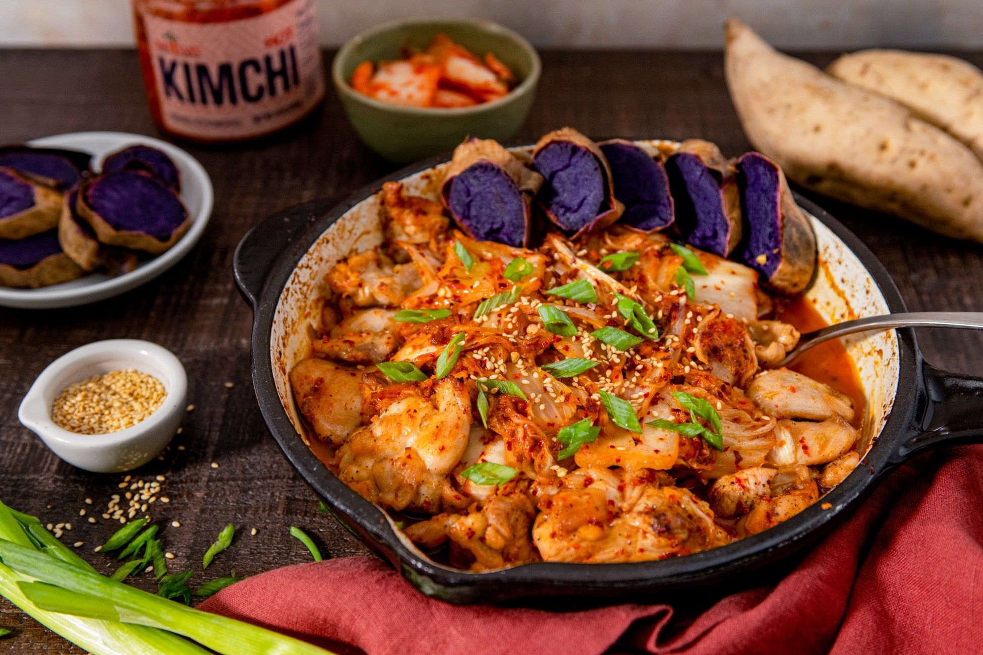 A bowl of spicy kimchi stew with purple sweet potatoes and garnishes.