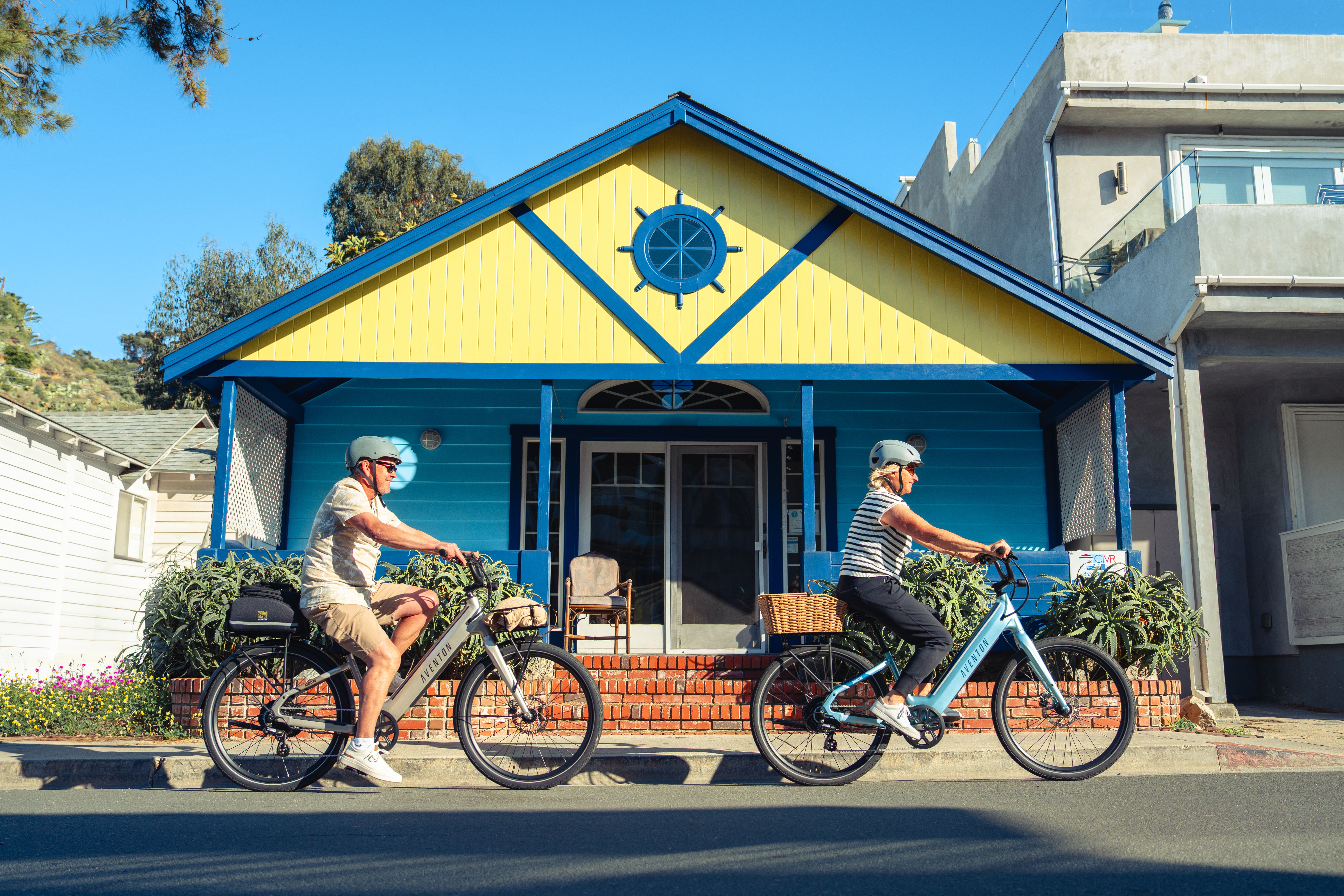 Two cyclists ride past a colorful house with a sun emblem.