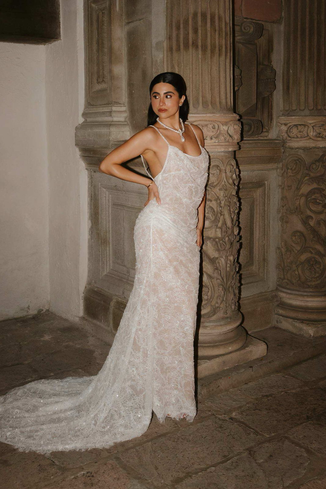 A woman in a white lace dress poses elegantly near ornate pillars.