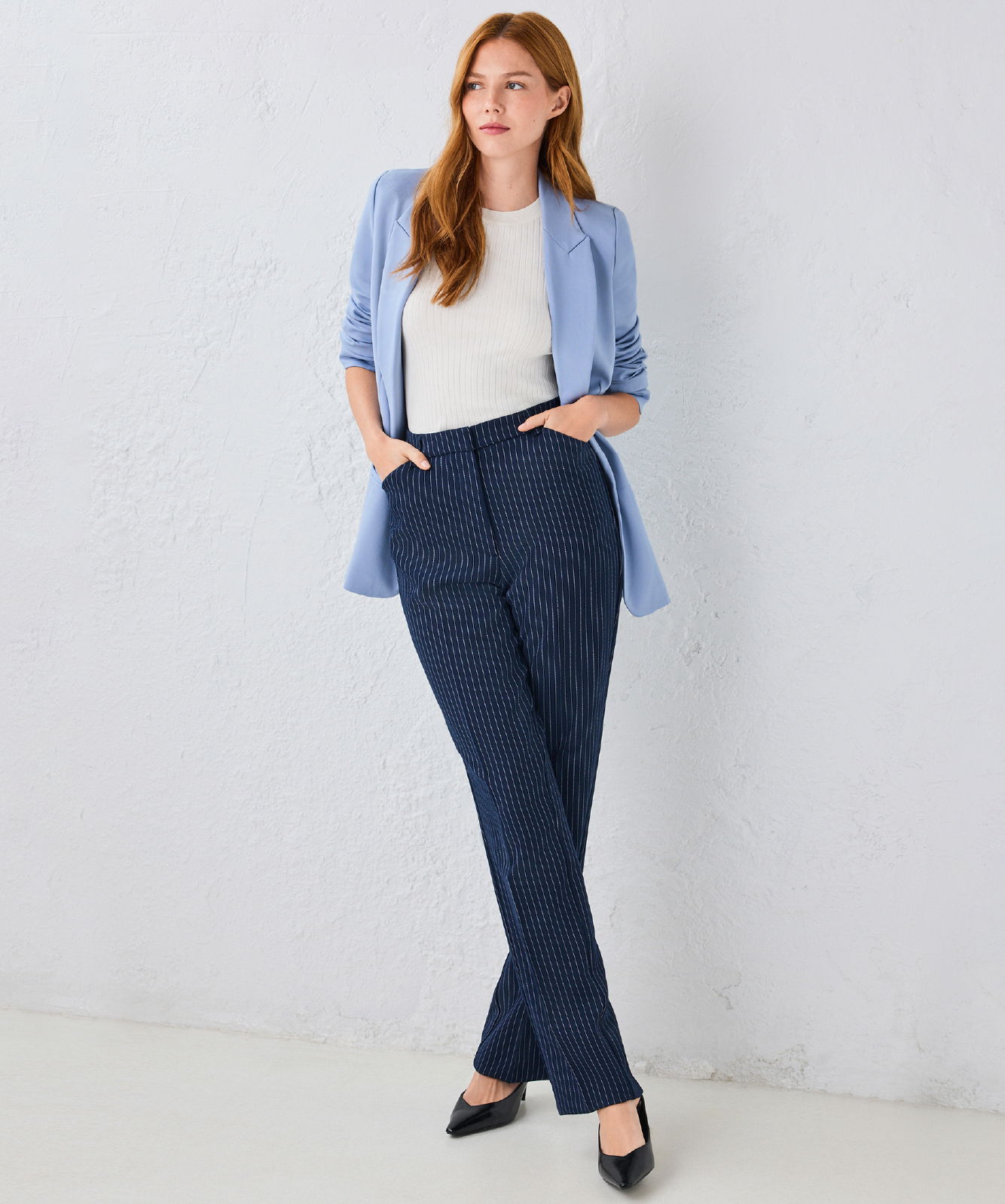 Woman in blue shirt and white top wearing dark pants, standing against a light background.