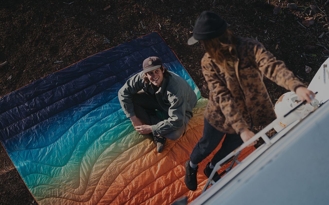Two people on a colorful quilt near a camper.